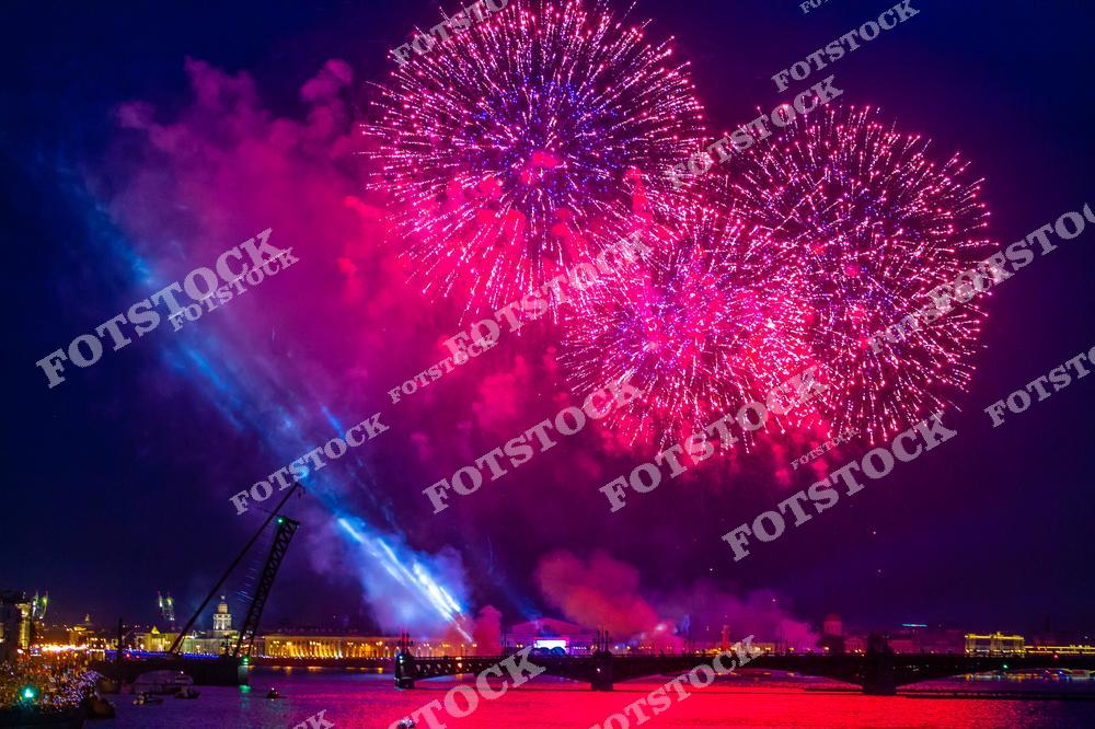 На фотографии изображён красочный фейерверк над рекой Невой в Санкт-Петербурге, в момент разведения одного из мостов. Праздничные огни освещают ночное небо, отражаясь в воде и создавая неповторимую атмосферу. На заднем плане видны исторические здания и подсвеченные набережные. Снимок передаёт торжественность и масштабность события.